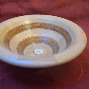 Può includere: Una ciotola di legno con un design circolare. La ciotola è realizzata in legno chiaro e scuro, con anelli concentrici di colori alternati. La ciotola è appoggiata su una superficie rossa.