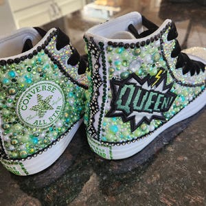 May include: Customized high-top sneakers with a white rubber sole. The canvas uppers are embellished with green and clear beads, rhinestones, and pearls. The design includes the word "QUEEN" in a comic book style and the Converse All Star logo.