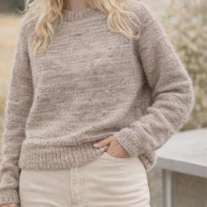 May include: A beige crew neck jumper with long sleeves. The jumper has a textured knit pattern and ribbed cuffs and hem. The person is wearing beige trousers and has their hand in the pocket of the trousers.
