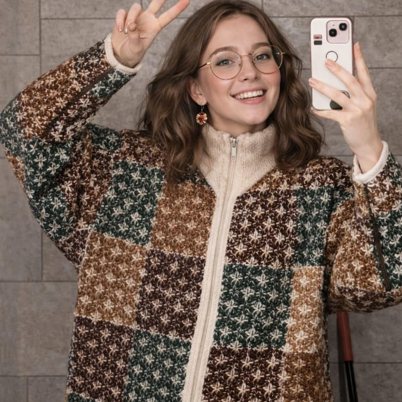 May include: A patchwork zip-up sweater in brown, green, and cream. The sweater has a high collar and a star pattern. The person in the image is wearing round glasses and holding a phone.