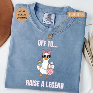 May include: Blue jean colored t-shirt with a graphic of a goose wearing sunglasses, a pink bow, and holding a coffee cup and a purse. The text reads "OFF TO... RAISE A LEGEND".