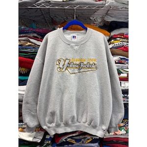 May include: A gray crewneck sweatshirt with the text "Georgia Tech Yellow Jackets" in yellow and navy blue. The sweatshirt has a classic fit and a ribbed neckline, cuffs, and hem. The Russell Athletic logo is visible on the upper chest.