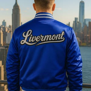 May include: Royal blue satin jacket with white and light blue striped trim. The word "Livermont" is embroidered on the back in white script with a black outline. The jacket is worn in front of a city skyline.