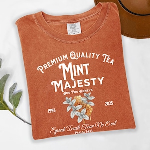 May include: An orange Comfort Colors t-shirt with the words "Premium Quality Tea Mint Majesty" in white script. The shirt features a floral design and the text "Speak Truth Fear No Evil" and the numbers 1993 and 2025.