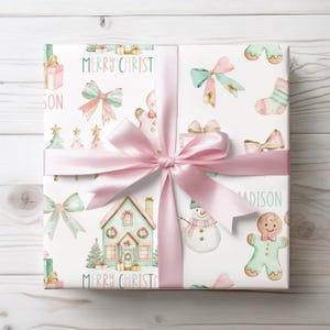 Holiday gift wrap with pastel bows, trees, and gingerbread cookies in pink and mint, shown mid-wrap with scissors and ribbon