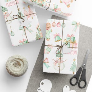 Holiday gift wrap with pastel bows, trees, and gingerbread cookies in pink and mint, shown mid-wrap with scissors and ribbon