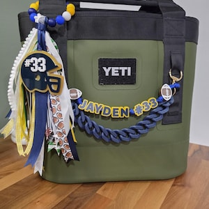 May include: An olive green YETI cooler bag with black straps, decorated with a football-themed accessory set. It features a blue and yellow beaded keychain with a number 33 helmet charm, ribbons, and a blue chain link strap reading JAYDEN #33.