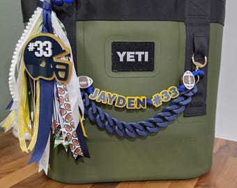 May include: An olive green YETI cooler bag with black straps, decorated with a football-themed accessory set. It features a blue and yellow beaded keychain with a number 33 helmet charm, ribbons, and a blue chain link strap reading JAYDEN #33.