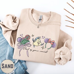 A beige sweatshirt with a graphic design featuring a cartoon character and the text "Part of your world" printed on it, along with a small charm. The sweatshirt is placed on top of a pair of blue jeans, and there are some brown twigs visible in the background.