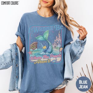 A woman wearing a blue t-shirt with a graphic design featuring a mermaid and a castle, along with the text "I Wanna Be Where The People Arent".