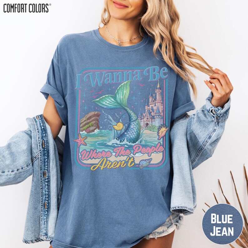 A woman wearing a blue t-shirt with a graphic design featuring a mermaid and a castle, along with the text "I Wanna Be Where The People Arent".