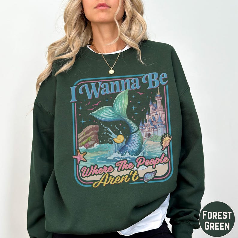 A woman wearing a green sweatshirt with a colorful graphic design featuring a mermaid, castle, and text.