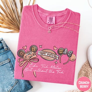 a pink t-shirt with a cartoon character design, a necklace, and a pair of jeans, all laid out on a white background.