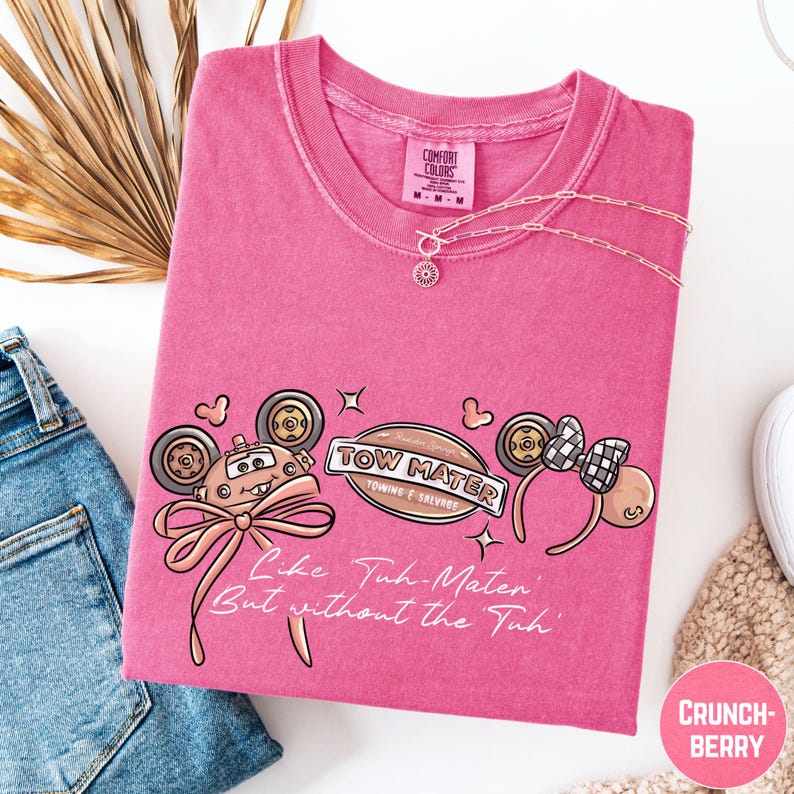 a pink t-shirt with a cartoon character design, a necklace, and a pair of jeans, all laid out on a white background.