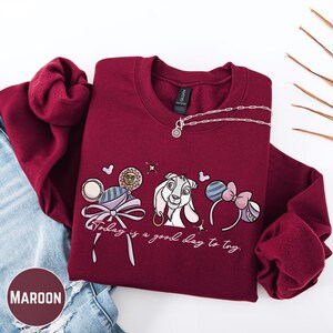 A maroon sweatshirt with a graphic design featuring a cartoon rabbit and the text "Today is a good day to try" is displayed on a white background.