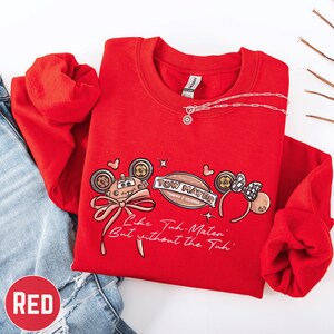 A red sweatshirt with a cartoon character design and text printed on it, along with a necklace and a pair of jeans.