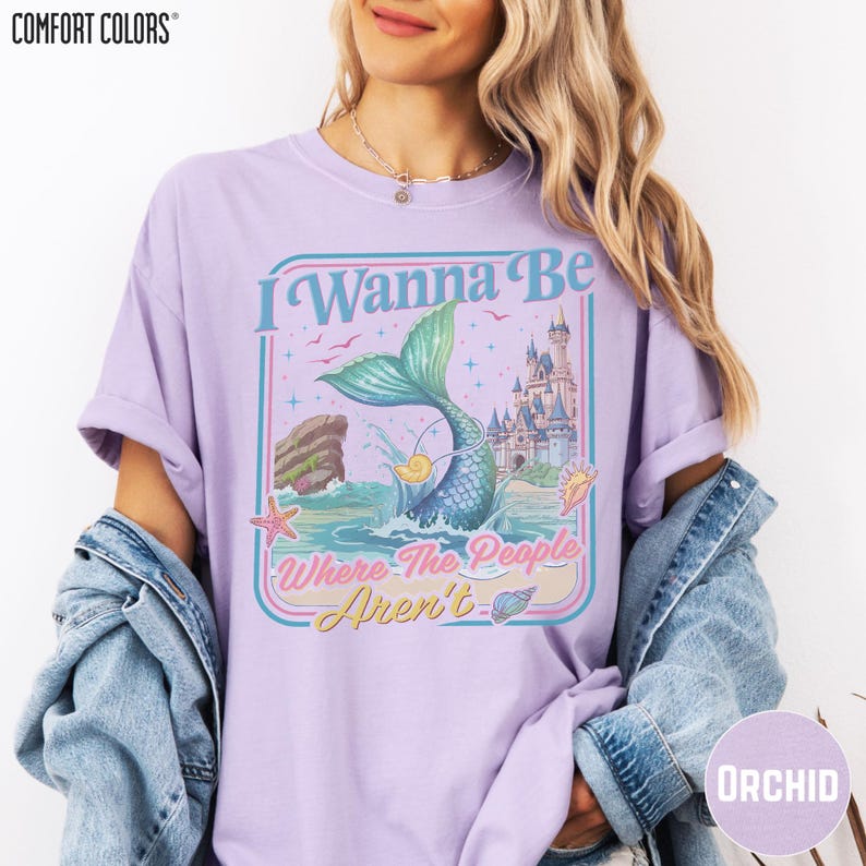 A woman wearing a purple t-shirt with a mermaid graphic and text, along with a denim jacket.