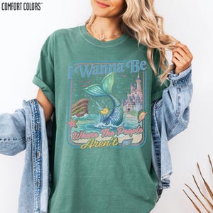 A woman wearing a green t-shirt with a graphic design featuring a mermaid and a castle, along with the text "I Wanna Be Where The People Arent".