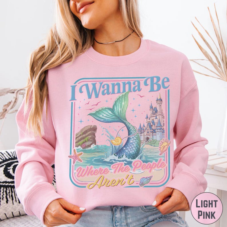A woman wearing a pink sweatshirt with a graphic design featuring a mermaid and a castle.