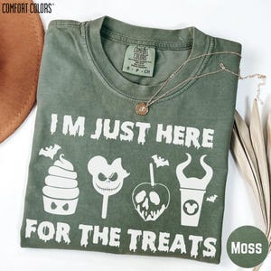 May include: A moss green Comfort Colors t-shirt featuring white graphic text that reads I'M JUST HERE FOR THE TREATS. The design includes illustrations of a cupcake, a skull lollipop, a poison apple, and a cup with horns, accented by bats.