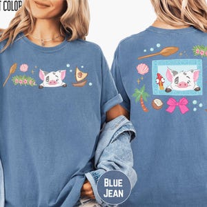 a woman wearing a blue t-shirt with a cartoon pig design and various colorful elements, such as a boat, flowers, and a bow.