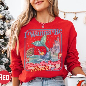 A woman wearing a red sweater with a mermaid graphic and text, standing in front of a Christmas tree.