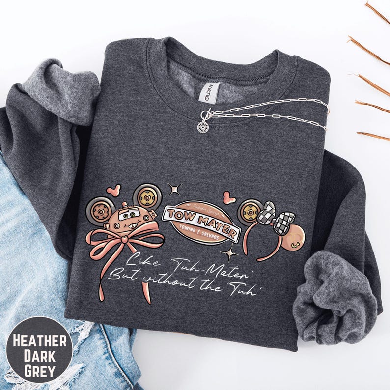 A gray sweatshirt with a graphic design featuring cartoon-like characters and text, along with a necklace and a pair of jeans.