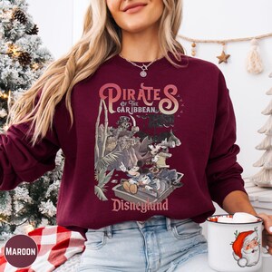 A woman wearing a burgundy sweatshirt with a graphic of Disney characters from the Pirates of the Caribbean ride at Disneyland.