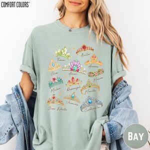 A woman wearing a light green t-shirt with a graphic design of various crowns and tiaras, along with the names of Disney princesses.