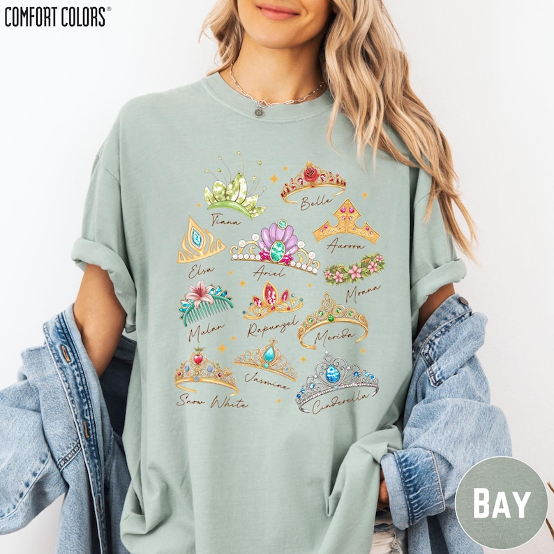 A woman wearing a light green t-shirt with a graphic design of various crowns and tiaras, along with the names of Disney princesses.
