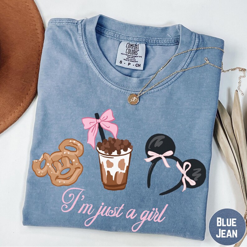 A blue t-shirt with a graphic design of a cup with a chocolate drink, a pair of Mickey Mouse ears, and a pink bow, along with the text "I'm just a girl".