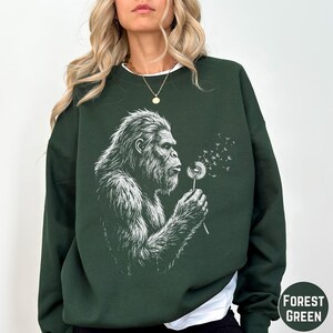 May include: A forest green long-sleeve sweatshirt featuring a white graphic illustration of a hairy creature blowing on a dandelion, with seeds floating away. The sweatshirt has a crew neckline and long sleeves, and the text Forest Green is visible.