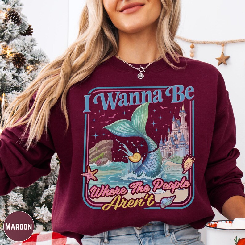 A woman wearing a burgundy sweatshirt with a graphic design featuring a mermaid and a castle.