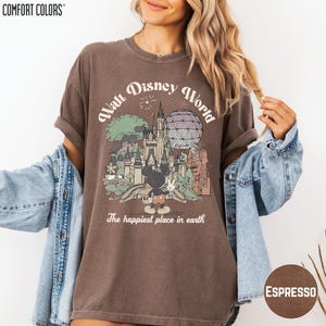 A woman wearing a brown t-shirt with a graphic of Disney World and the text "The happiest place in earth" is posing with her arms outstretched.