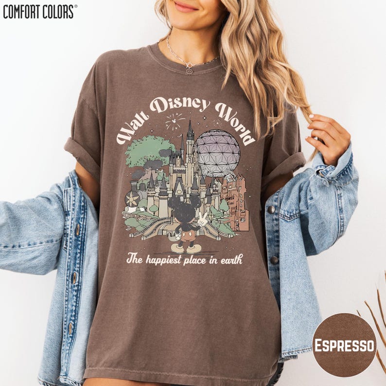 A woman wearing a brown t-shirt with a graphic of Disney World and the text "The happiest place in earth" is posing with her arms outstretched.