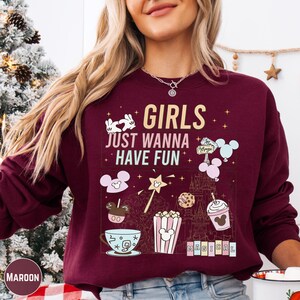 A woman wearing a burgundy sweatshirt with a graphic design featuring various Disney-themed items and text.