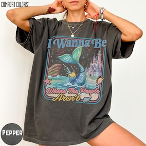 A person wearing a black t-shirt with a graphic design featuring a mermaid and the text "I Wanna Be Where The People Aren't".