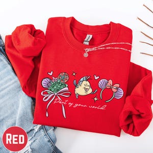 A red sweater with a cartoon character design and the text "Part of your world" printed on it, along with a necklace and a pair of jeans.