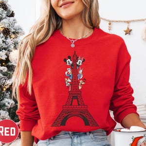 A woman wearing a red sweater with a graphic of the Eiffel Tower and Disney characters on it, standing in front of a Christmas tree.