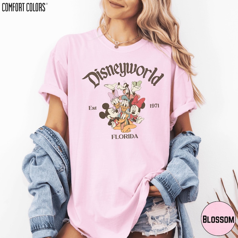 A woman wearing a pink t-shirt with a Disneyworld graphic and text, along with a denim jacket, posing against a white background.