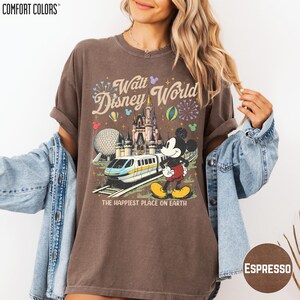 A woman wearing a brown t-shirt with a Disney World graphic and text, along with a denim jacket, posing against a white background.