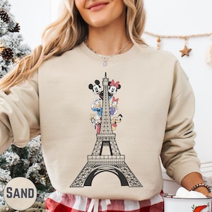A woman wearing a beige sweater with a graphic of the Eiffel Tower and Disney characters on it.