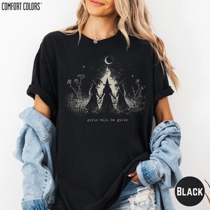 May include: A black short-sleeve t-shirt featuring a white graphic print of three figures in pointed hats standing in a field under a crescent moon. The text "girls will be girls" is printed below the figures. The brand Comfort Colors is visible.
