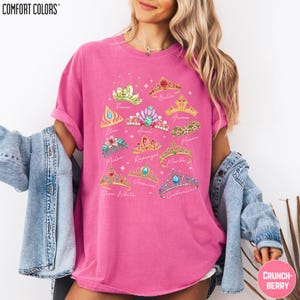 A woman wearing a pink t-shirt with a graphic design of various crowns and tiaras, along with the names of the characters associated with them. She is also wearing a denim jacket and has her arms around her shoulders.