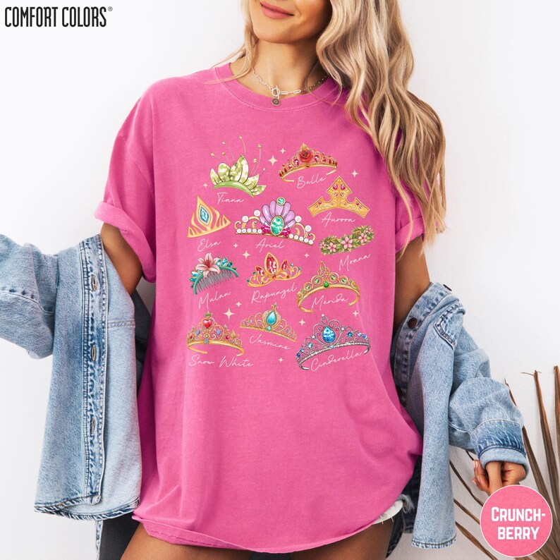 A woman wearing a pink t-shirt with a graphic design of various crowns and tiaras, along with the names of the characters associated with them. She is also wearing a denim jacket and has her arms around her shoulders.