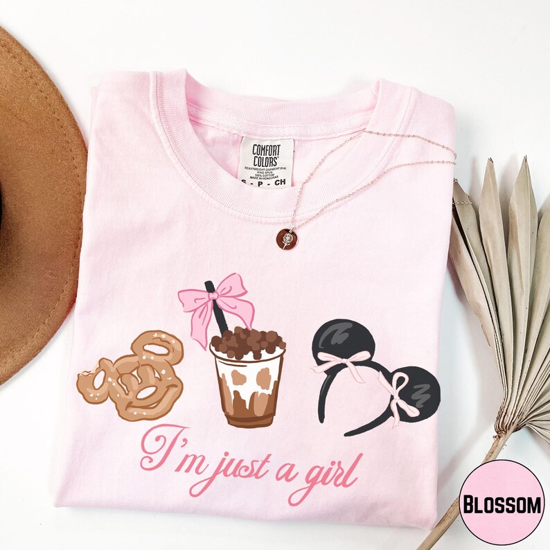 a pink t-shirt with a graphic design of a cup with a chocolate drink, a pair of Mickey Mouse ears, and a cookie. The text on the t-shirt says "I'm just a girl".