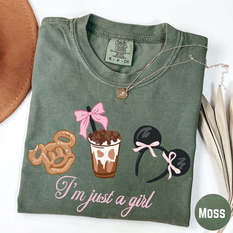 a green t-shirt with a graphic design of a cup with a pink ribbon, a pretzel, and a pair of earrings, along with the text "I'm just a girl".