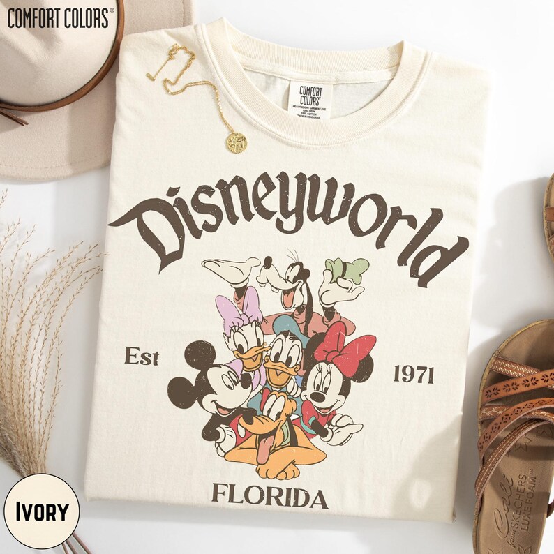 a white t-shirt with a Disney World graphic, including Mickey Mouse, Minnie Mouse, Goofy, Donald Duck, and Pluto, along with the text "Disneyworld" and "Est. 1971 FLORIDA". The t-shirt is displayed on a white background, and there are some accessories, such as a hat and sandals, visible in the image.