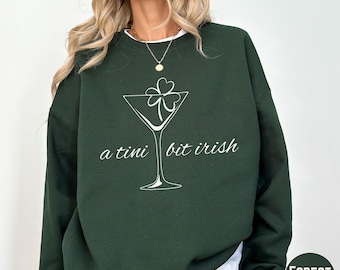 May include: Forest green sweatshirt with a white graphic of a martini glass with a shamrock and the text "a tini bit irish". The sweatshirt has a white crew neck and is a solid color.