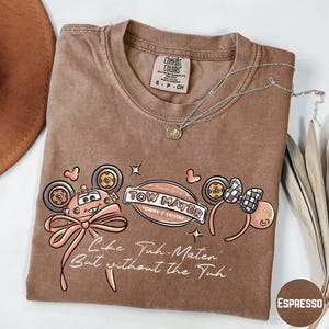 a brown t-shirt with a cartoon character design, a necklace, and some decorative elements.